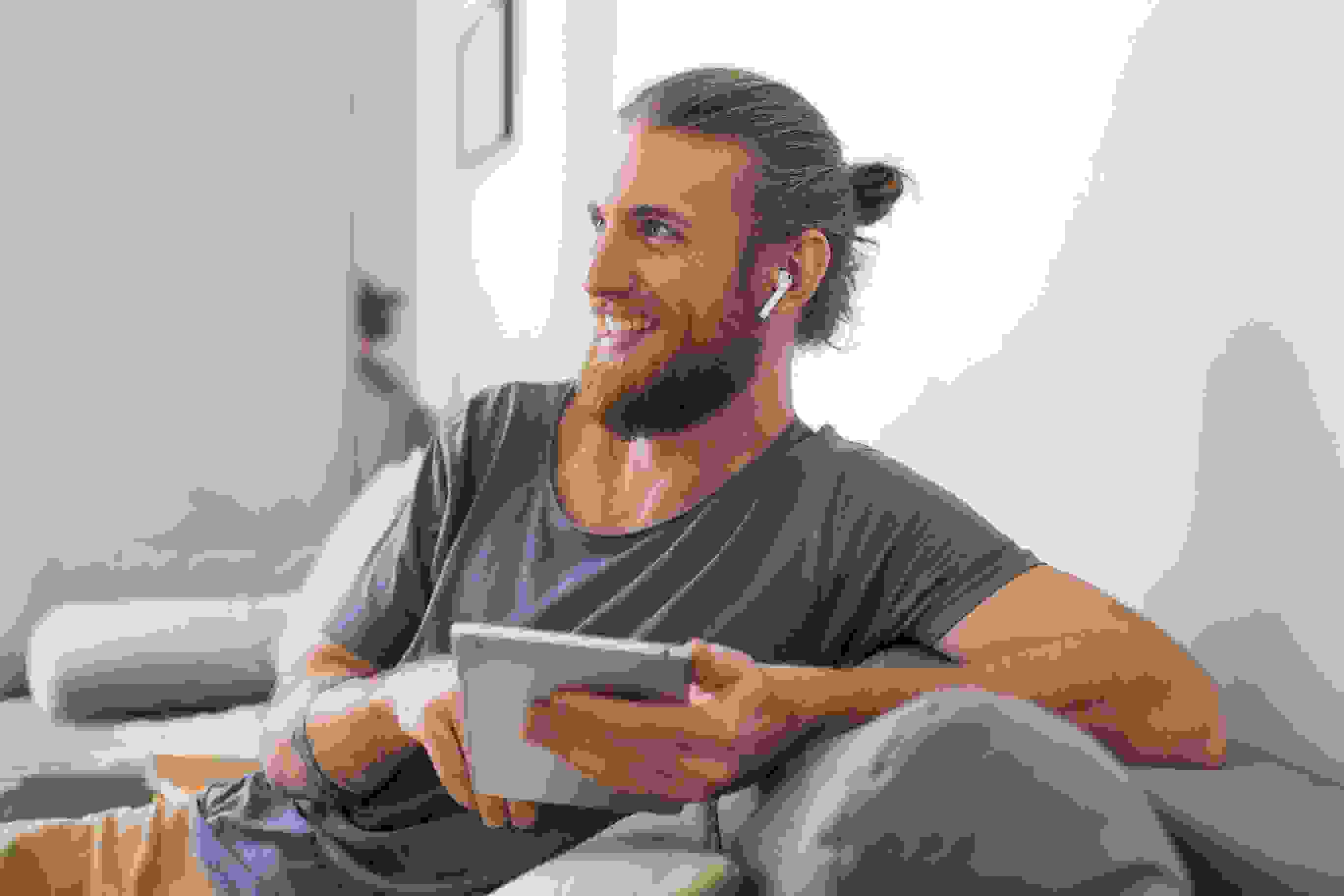 portrait-young-man-sitting-gray-sofa-with-earphones-tablet-hands-happily-looking-aside-home